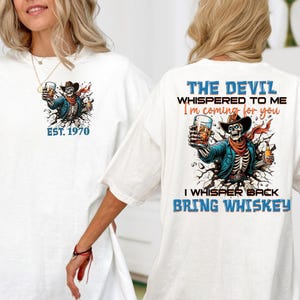 May include: White t-shirt featuring a skeleton in a cowboy hat, holding a whiskey glass. The front has "EST. 1970". The back reads "THE DEVIL WHISPERED TO ME I'm coming for you I WHISPER BACK BRING WHISKEY".