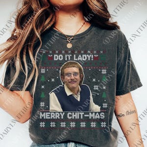 May include: Dark gray t-shirt with a festive design. The shirt features the text "DO IT LADY!" above an image of a man wearing glasses and a sweater vest. Below the image is the text "MERRY CHIT-MAS" with snowflake and Christmas tree graphics.
