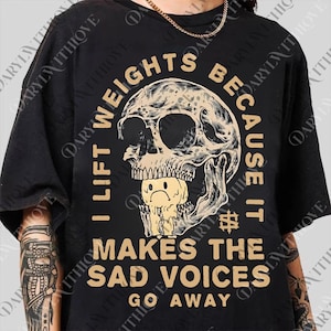 May include: Black t-shirt with a distressed skull graphic and the text "I Lift Weights Because It Makes The Sad Voices Go Away." The skull has a sad face inside its mouth. The text and skull are in a tan color.
