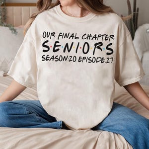 May include: A cream-colored short-sleeve t-shirt featuring black text that reads Our Final Chapter Seniors Season: 20 Episode: 27. The word Seniors is written in a bold font with small colored dots between the letters in blue, red, and yellow.