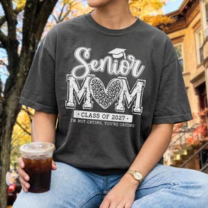 May include: A charcoal gray short-sleeve t-shirt featuring white screen-printed text that reads Senior MOM with a leopard print heart in the center, a graduation cap icon, and the text CLASS OF 2027 I'M NOT CRYING, YOU'RE CRYING below the heart.