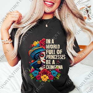 Peut inclure: T-shirt noir avec un graphisme coloré d'une femme avec un maquillage de tête de mort, un chapeau fleuri et le texte "In a world full of princesses be a chingona."