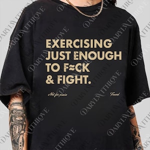 Könnte beinhalten: Schwarzes T-Shirt mit goldenem Text: "EXERCISING JUST ENOUGH TO F**K & FIGHT." Darunter stehen die Worte "Not for pussies" und "Frosed." Das Shirt hat kurze Ärmel und eine lockere Passform.