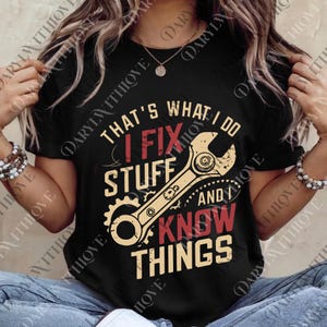 以下が含まれることがあります： 「THAT'S WHAT I DO, I FIX STUFF AND I KNOW THINGS」という赤とベージュの文字がプリントされた黒いTシャツ。レンチと歯車のグラフィックがテキストの下に表示されています。半袖のシャツです。