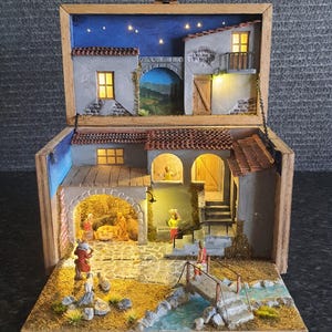 Könnte beinhalten: Eine Miniatur-Krippenszene in einer offenen Holzschachtel mit beleuchteten Gebäuden mit Terrakottadächern, einem gepflasterten Innenhof, einer kleinen Brücke über einen blauen Bach und mehreren kleinen Figuren in einem Dorf-Szenario.