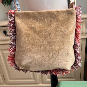 May include: A brown suede crossbody bag with a fringe trim in red, white, and burgundy. The bag has a long adjustable strap.