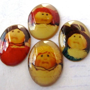 Könnte beinhalten: Vier ovale Cabochons mit Bildern von Cabbage Patch Kids. Jeder Cabochon hat ein glänzendes, transparentes Finish. Die Bilder zeigen die Puppen mit unterschiedlichen Haarfarben und Outfits, vor einem gelben Hintergrund.