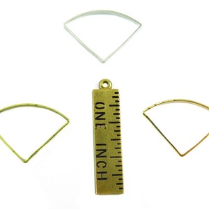 May include: An assortment of metal fan-shaped jewelry findings in silver, gold, and brass tones. A brass ruler with the text "ONE INCH" is also present, indicating the size of the findings. These components are ideal for crafting earrings, pendants, or other jewelry.