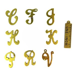 May include: A collection of vintage brass letter charms in a variety of cursive fonts. The letters include C, F, G, K, P, R, and V. A brass ruler with the text "ONE INCH" is also included. The charms are on a white background.