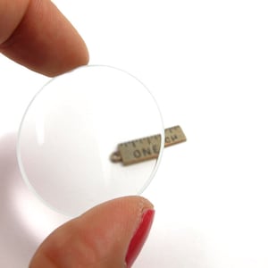 May include: A clear, circular magnifying lens is held between two fingers. A small, rectangular charm with the text "ONE INCH" is visible in the background. The background is white.