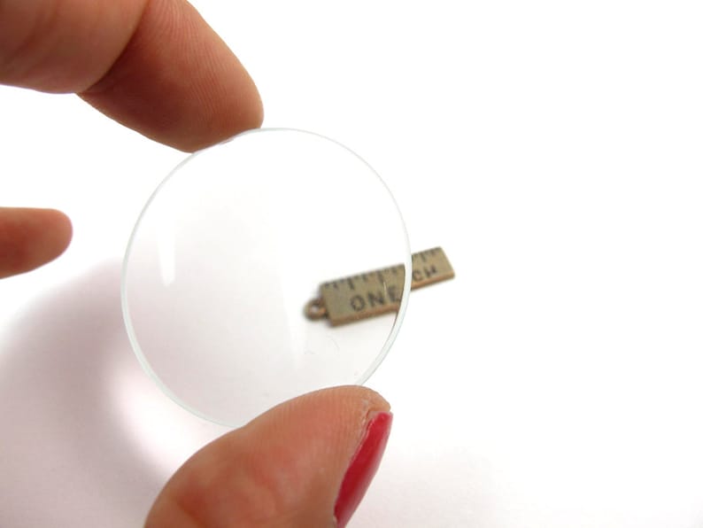 May include: A clear, circular magnifying lens is held between two fingers. A small, rectangular charm with the text "ONE INCH" is visible in the background. The background is white.