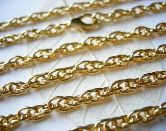 May include: Close-up of several strands of gold-colored chain. The chains are linked together with a wheat-style pattern. A gold-colored clasp is visible in the center. The chains are arranged diagonally across a white surface.