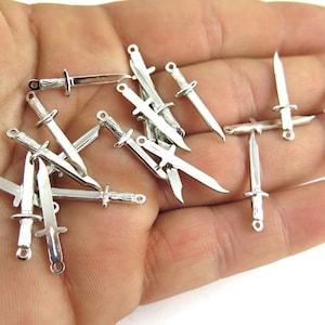 May include: A collection of silver-toned metal knife charms. The charms are in the shape of a knife with a pointed tip and a handle. The charms are scattered on a hand.