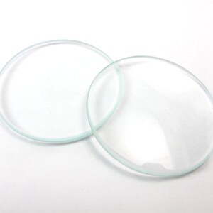 May include: Two clear, circular glass lenses are displayed on a white surface. One lens is a complete circle, the other is slightly overlapping, appearing as a flattened oval. The glass is transparent with a slight greenish tint.