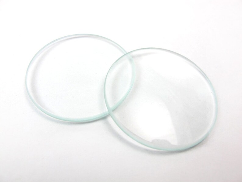 May include: Two clear, circular glass lenses are displayed on a white surface. One lens is a complete circle, the other is slightly overlapping, appearing as a flattened oval. The glass is transparent with a slight greenish tint.