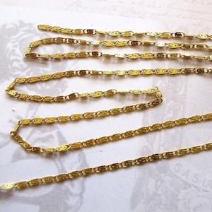 May include: A close-up of a gold chain with a decorative, patterned design. The chain is made of small, linked pieces that are shaped like a letter 'S'.