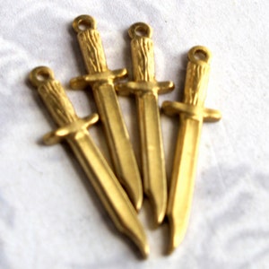 May include: Four gold-colored metal dagger charms with a detailed design. The charms are arranged in a cluster, with the points of the daggers facing outward.