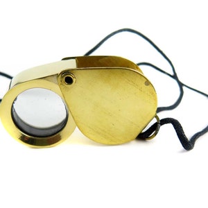 May include: A brass and ivory-colored magnifying glass with a black cord. The lens is circular and set in a gold-colored frame. The magnifying glass is open, revealing the lens, and the cord is looped.