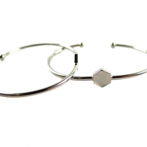 May include: Two silver-colored, open-ended cuff bracelets. One bracelet has a simple design, while the other features a small, geometric, hexagonal charm. The bracelets are set against a white background.