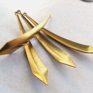 May include: Four gold-colored, elongated metal pendants with pointed ends and small loops at the top. The pendants have a slightly curved shape and a matte finish, lying on a light-colored surface. The design is geometric and minimalist.