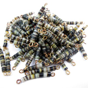 May include: A pile of small, cylindrical beads with a striped pattern in shades of black, brown, and cream. Each bead has a gold-colored loop at each end, suggesting they are designed for jewelry making or crafts.