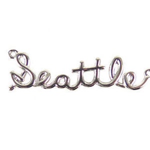 Könnte beinhalten: Ein silberfarbener Draht-Wort-Anhänger, der "Seattle" in einer Schreibschrift darstellt. Der Anhänger hat an jedem Ende kleine Schlaufen, was darauf hindeutet, dass er als Anhänger oder Charm für Schmuck oder Bastelarbeiten verwendet werden kann.