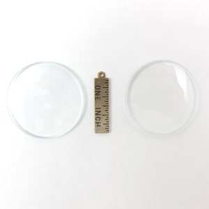 May include: Two clear, circular glass lenses with a brass ruler that reads "ONE INCH". The lenses are transparent and appear to be convex. The ruler is vertically oriented between the two lenses.