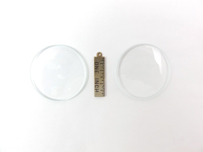 May include: Two clear, circular glass lenses with a brass ruler that reads "ONE INCH". The lenses are transparent and appear to be convex. The ruler is vertically oriented between the two lenses.