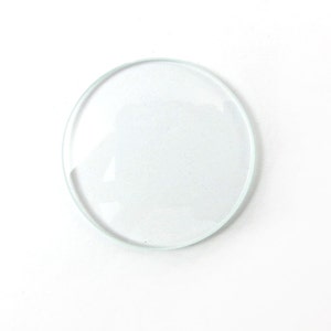 May include: A transparent, circular glass lens with a raised edge. The lens is clear and appears to be a single piece of glass. The background is plain white.