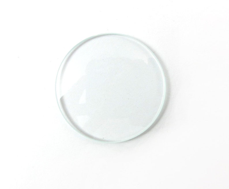 May include: A transparent, circular glass lens with a raised edge. The lens is clear and appears to be a single piece of glass. The background is plain white.