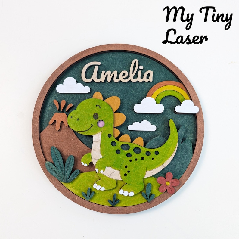 May include: A round, layered wooden sign featuring a green dinosaur, rainbow, clouds, and the name "Amelia". The sign has a brown border and includes the text "My Tiny Laser". The dinosaur has yellow accents and black spots.