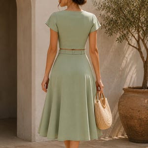 May include: A light green two-piece outfit featuring a short-sleeved crop top and a midi skirt with a matching belt. A woven straw handbag is held, and the outfit is paired with beige heels. The background includes an olive tree in a large pot.