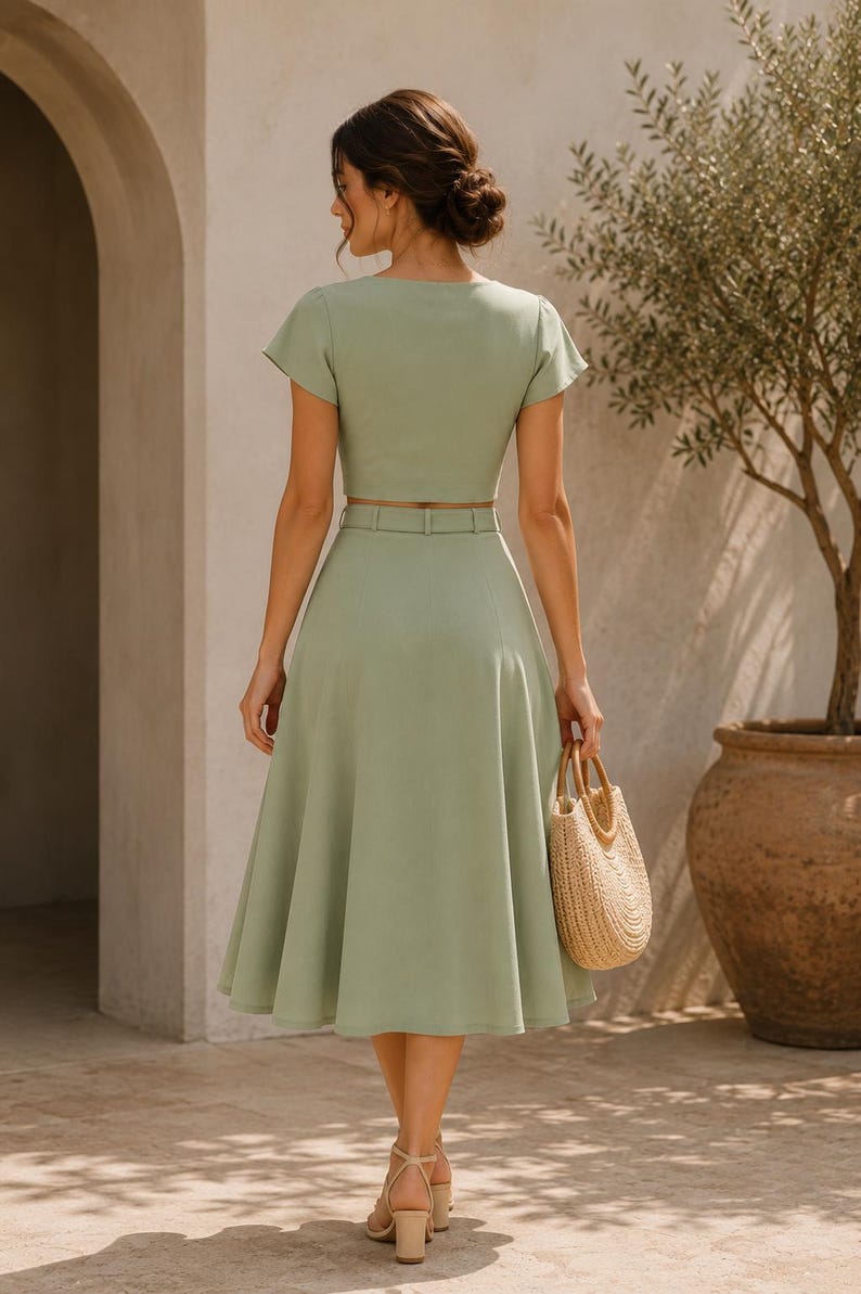 May include: A light green two-piece outfit featuring a short-sleeved crop top and a midi skirt with a matching belt. A woven straw handbag is held, and the outfit is paired with beige heels. The background includes an olive tree in a large pot.