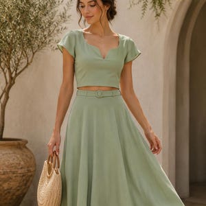 May include: A sage green two-piece outfit featuring a crop top with a scalloped neckline and a matching midi skirt. The outfit includes a thin belt with a circular buckle. A woven straw handbag with wooden handles completes the look.