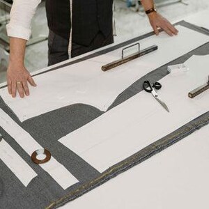 May include: A tailor's workspace with white paper patterns laid on gray fabric. Scissors, metal weights, and wooden tools are arranged on the cutting table. A person's hands are visible, suggesting the process of garment creation.