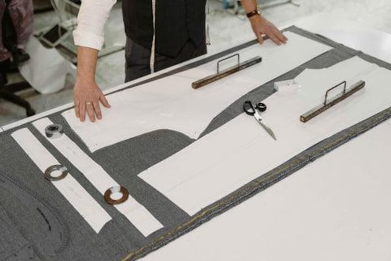 May include: A tailor's workspace with white paper patterns laid on gray fabric. Scissors, metal weights, and wooden tools are arranged on the cutting table. A person's hands are visible, suggesting the process of garment creation.