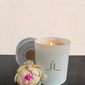 May include: White candle with a gold logo and a lit wick. The candle is in a glass jar with a white lid. A pink peony flower is in the foreground.