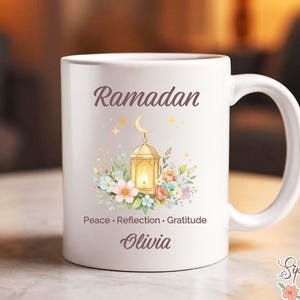May include: White ceramic mug with the word "Ramadan" in a script font. The mug features a gold lantern design with a crescent moon and floral accents. The words "Peace, Reflection, Gratitude" and a name are also printed on the mug.