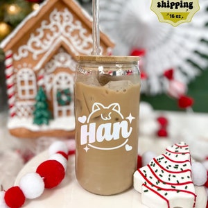 May include: A clear glass jar with a bamboo lid and a straw, filled with iced coffee. The jar has a white decal with the name "Han" and a cartoon cat design. The jar is sitting on a white surface with red and white pom-poms and a white and red candy cane Christmas tree.