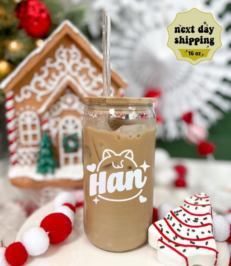 May include: A clear glass jar with a bamboo lid and a straw, filled with iced coffee. The jar has a white decal with the name "Han" and a cartoon cat design. The jar is sitting on a white surface with red and white pom-poms and a white and red candy cane Christmas tree.