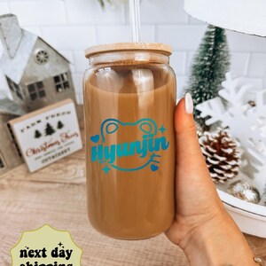 May include: A clear glass reusable 16 ounce tumbler with a bamboo lid and a blue and green frog graphic with the text "Hyunjin".