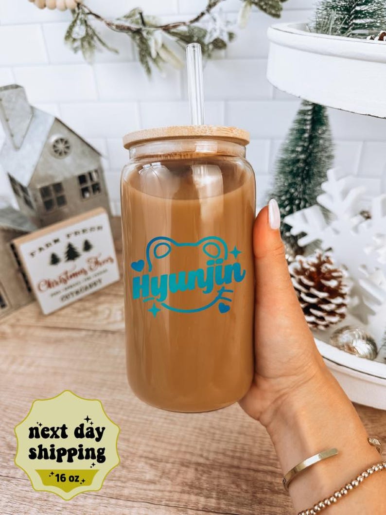 May include: A clear glass reusable 16 ounce tumbler with a bamboo lid and a blue and green frog graphic with the text "Hyunjin".