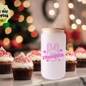 May include: White reusable tumbler with a pink bunny and the text "Changbin" in pink. The tumbler has a bamboo lid and a clear straw. The tumbler is sitting on a table with cupcakes in the background.