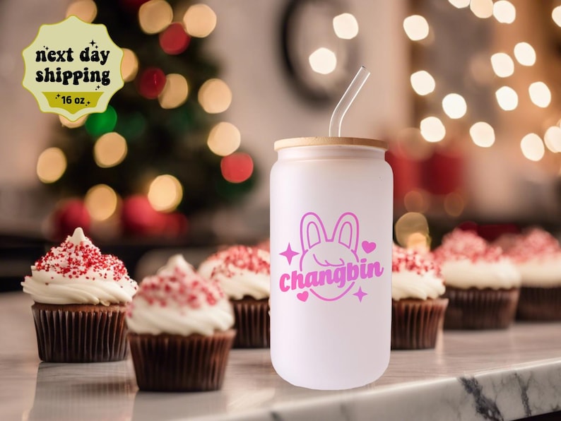 May include: White reusable tumbler with a pink bunny and the text "Changbin" in pink. The tumbler has a bamboo lid and a clear straw. The tumbler is sitting on a table with cupcakes in the background.