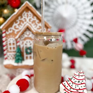 May include: A glass jar with a wooden lid filled with iced coffee and a clear glass straw. The jar is sitting on a white surface with red and white pom-poms and a white gingerbread cookie shaped like a Christmas tree.