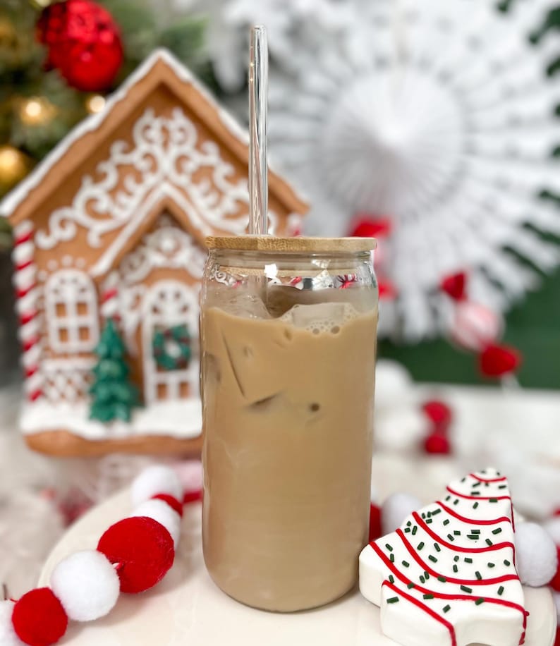 May include: A glass jar with a wooden lid filled with iced coffee and a clear glass straw. The jar is sitting on a white surface with red and white pom-poms and a white gingerbread cookie shaped like a Christmas tree.