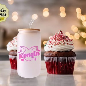 May include: A pink and white 16 oz. reusable tumbler with a bamboo lid and a pink cat design with the name "Jeongjin" in pink letters. The tumbler is sitting on a white surface next to two chocolate cupcakes with red and white frosting.