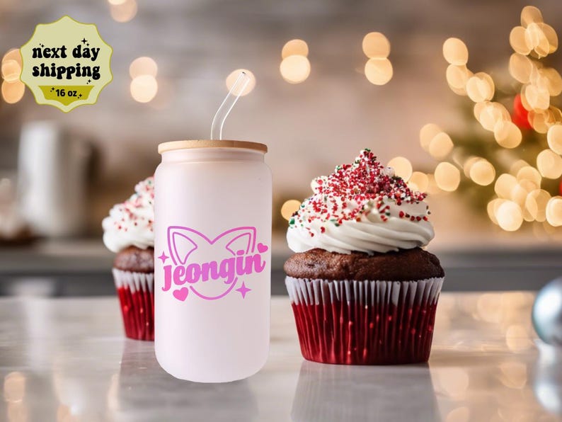 May include: A pink and white 16 oz. reusable tumbler with a bamboo lid and a pink cat design with the name "Jeongjin" in pink letters. The tumbler is sitting on a white surface next to two chocolate cupcakes with red and white frosting.