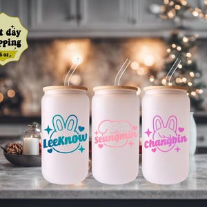 May include: Three 16 oz. reusable tumblers with lids and straws. Each tumbler features a different pink and white cartoon bunny with a name underneath: Lee Know, Seungmin, and Changbin.