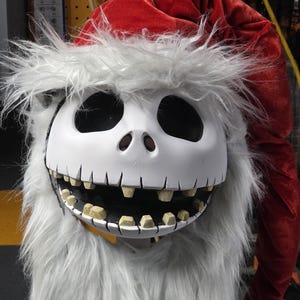 Puede incluir: Una máscara blanca con grandes cuencas de ojos negros y una amplia sonrisa con dientes amarillos. La máscara está rodeada de larga y gris piel sintética y coronada con un gorro de Papá Noel rojo. La máscara es un personaje de la película "Pesadilla antes de Navidad".